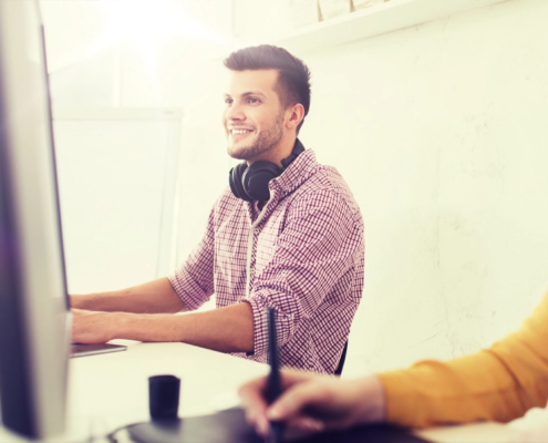A smiling man wearing headphones sits at a computer, taking notes.